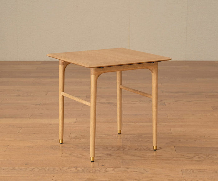The Hendrick End Table - White Oak, a mid-century design crafted from solid white oak with slim, rounded legs and elegant brass caps, stands on a matching wooden floor against a plain beige wall.