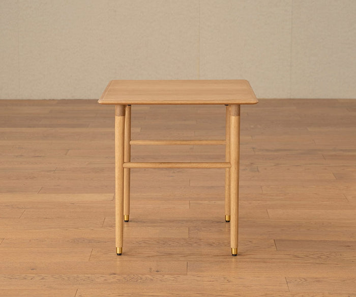 The Hendrick End Table – White Oak stands on a wooden floor against a plain, beige wall. This compact, light wood square table features rounded edges and four legs finished with elegant brass caps. Inspired by classic mid-century design, it is crafted from solid white oak for lasting beauty and durability.