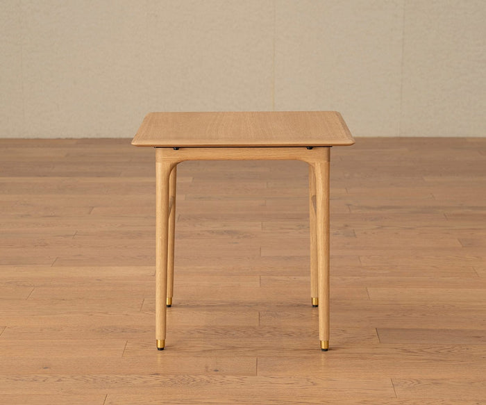 The Hendrick End Table - White Oak, featuring a solid white oak construction with four tapered legs and brass caps, stands on a wooden floor against a plain beige wall.