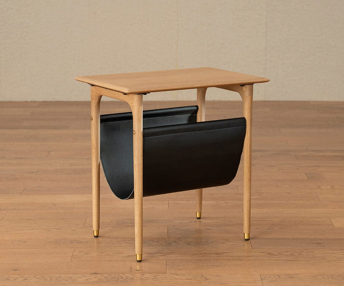 The Hendrick End Table With Hanging Shelf - White Oak, a compact mid-century design crafted from solid white oak, features a hanging black leather shelf below for storing magazines or newspapers. Positioned on a wooden floor against a beige wall, its legs are accented with subtle brass caps that lend an elegant touch to this stylish piece.