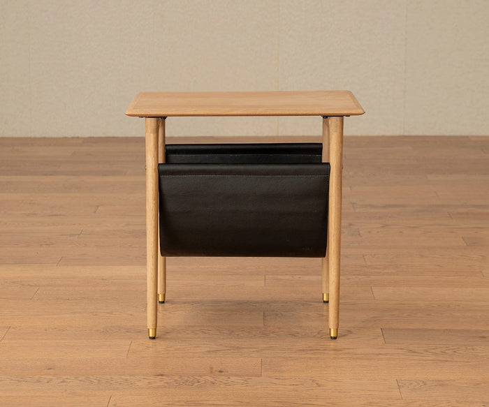 The Hendrick End Table With Hanging Shelf – White Oak features solid white oak legs with elegant brass caps, a matching rectangular top, and a black leather or faux-leather magazine holder suspended underneath. This small mid-century end table is showcased on a light wood floor against a neutral wall.