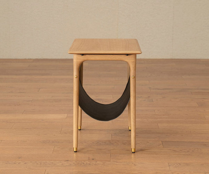 The Hendrick End Table With Hanging Shelf - White Oak is a small, mid-century style table with four legs featuring brass caps and gold-colored accents, as well as a black fabric sling shelf underneath. It is shown placed on a light wooden floor against a beige wall.