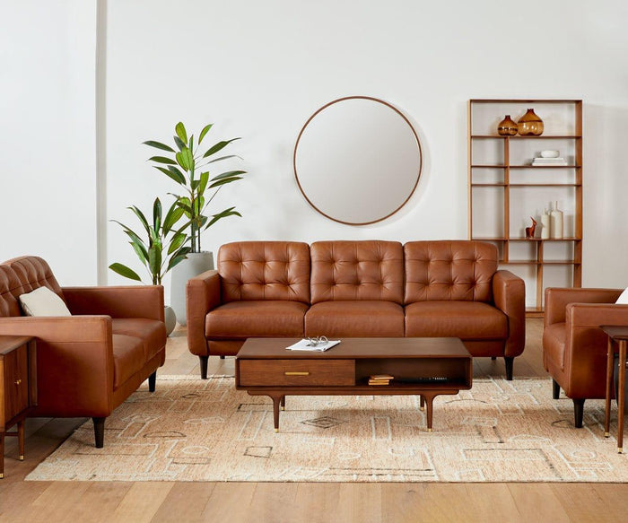 A modern living room with brown leather sofas, a wooden coffee table, the Hendrick End Table with Hanging Shelf featuring sleek brass leg caps, a tall plant, a round mirror, and a shelving unit with decor—all set on a light patterned rug and wood flooring.