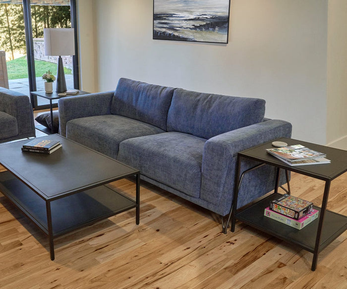 A contemporary living room features a blue couch, black coffee table topped with a plant for freshness, and the Heroy End Table—its matte black finish seamlessly blending into the industrial decor. The Heroy End Table is styled with books and a mouse. Set on wooden flooring, the space also has a large window and a painting displayed on the wall.