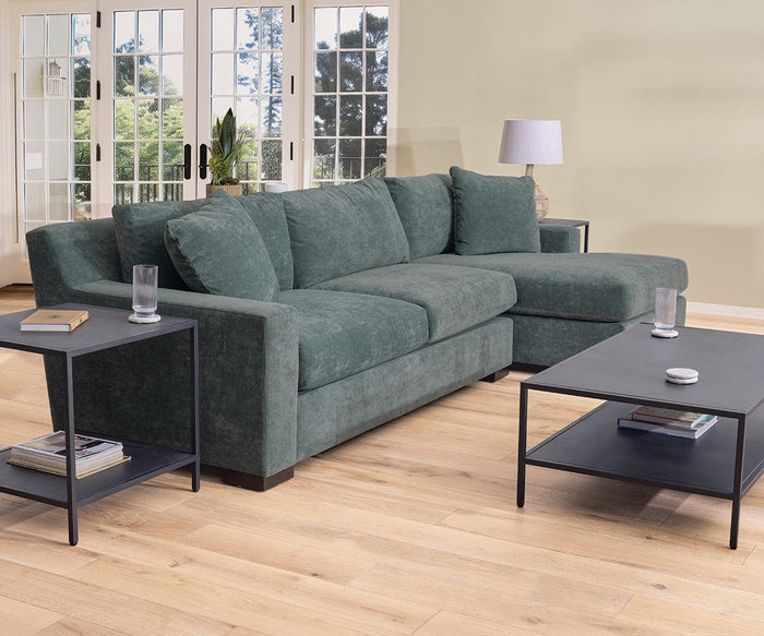 A green sectional sofa with chaise is arranged on a light wood floor. Glasses of water and books are placed on the matte black Heroy End Table, which is paired with matching coffee tables. Large windows in the background allow natural light to flood the room, beautifully enhancing the space’s industrial decor.