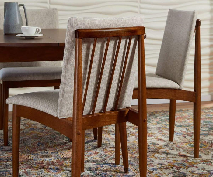 Two Holfred Dining Chairs, featuring light upholstered cushions, are arranged around a dark wooden table to create a Scandi-inspired look. On the tabletop rest a cup and a metal pitcher, while a vibrant patterned rug and a wavy textured wall serve as the backdrop.