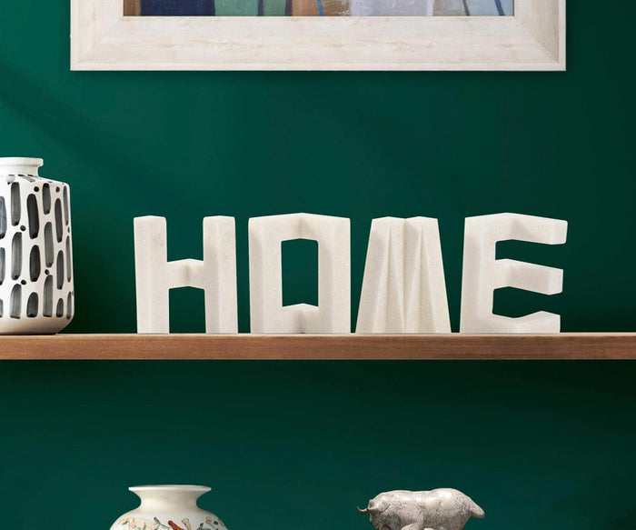 The Home Sculpture, a set of white decorative letters spelling HOME, sits on a wooden shelf against a dark green wall. The display is complemented by a patterned vase, a ceramic figure, and a painting, adding modern touches to the decor.