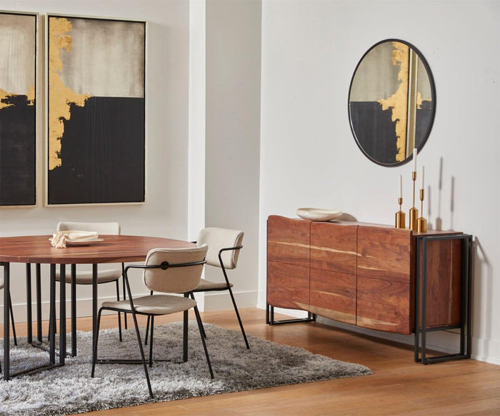 Modern dining room with a wooden table, four beige chairs, a wooden sideboard, a Janelle 36″ Round Mirror featuring a lacquered brass finish, abstract wall art, gold vases, and a gray rug on a wooden floor.