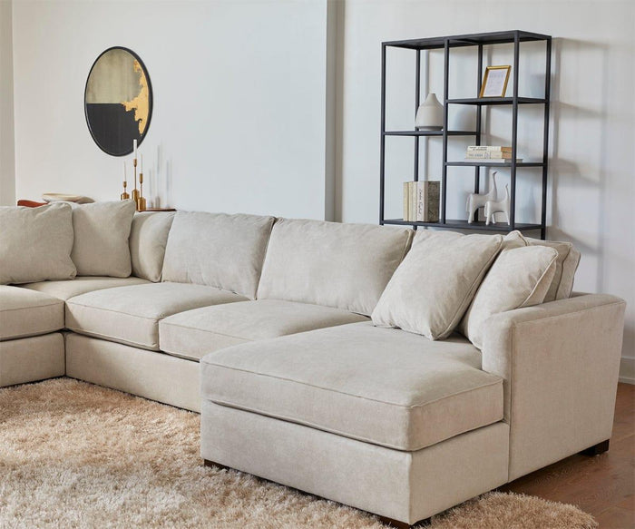 A beige sectional sofa sits on a light shaggy rug in the living room. Behind it, there’s a modern black metal shelf with books and decor, and the Janelle 36″ Round Mirror with a lacquered brass finish is mounted on the white wall.