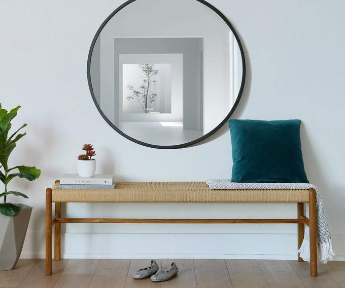 A modern wall mirror with a lacquered brass finish—the Janelle 36″ Round Mirror—hangs above a wooden bench adorned with a teal pillow, white book, and small plant. Shoes rest on the floor below, while a potted plant sits to the left. The Janelle 36″ Round Mirror reflects a bright hallway, adding elegance to the space.