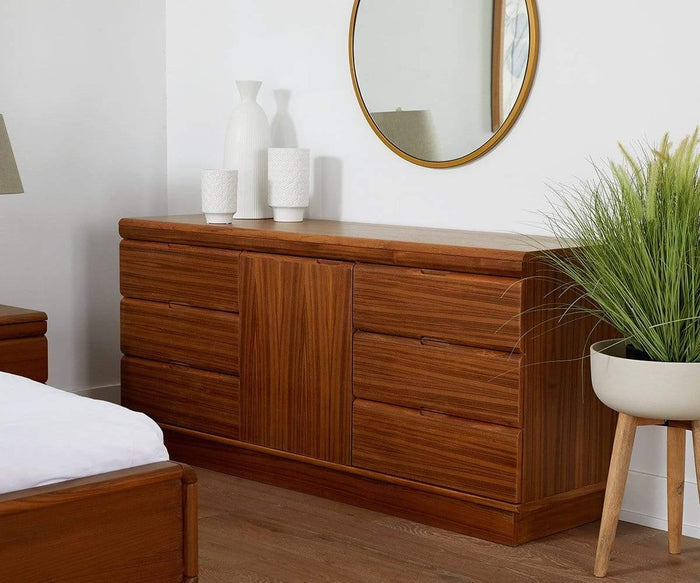 A wooden dresser with the Janelle 36″ Round Mirror—a modern wall mirror with a lacquered brass finish—hung above it, complemented by white decorative vases, a lamp, and a potted plant beside it in a contemporary bedroom setting.