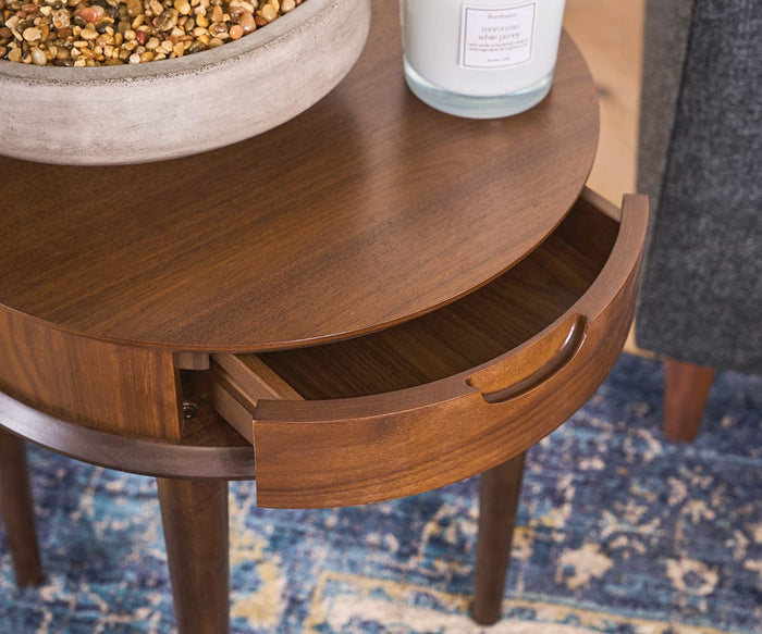 The Juneau End Table, featuring a handleless drawer partially open and topped with a potted plant and a candle, sits on a blue patterned rug next to a gray upholstered chair—an ideal living room accent that brings subtle mid-century charm to your space.