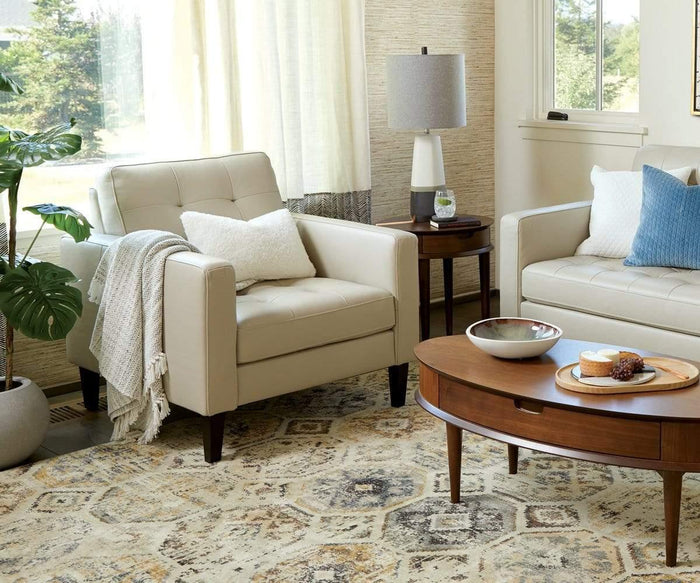A cozy living room features a cream armchair, sofa with pillows, a wooden coffee table with decor, and the stylish Juneau End Table beside the seating area. A potted plant, lamp, and patterned rug complete the inviting space as sunlight streams through a window with sheer curtains.