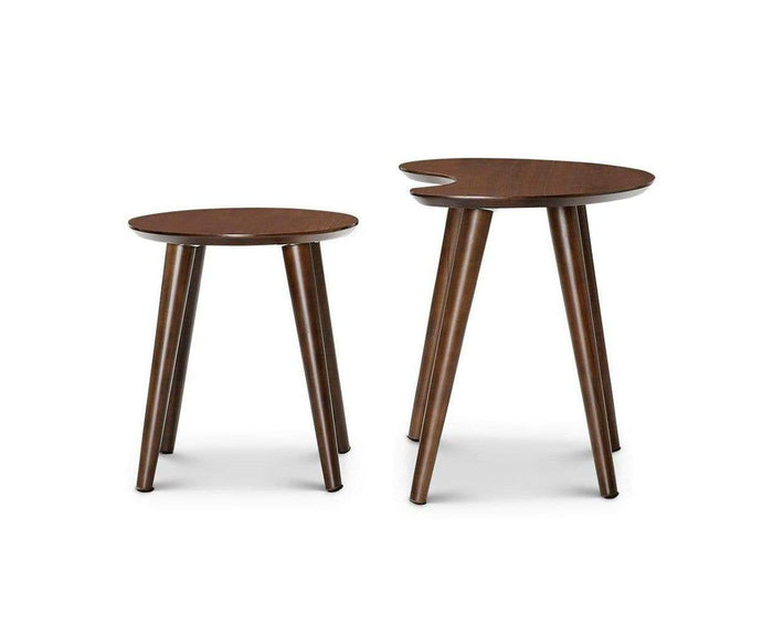The Juneau Nest Of Tables in a rich walnut color features two wooden side tables with round tops and three angled legs, channeling mid-century modern style. One table stands slightly taller with a distinctive curved cutout on its surface. Together, these dark walnut-finished tables create an elegant nesting set that complements any living space, shown here against a white background.