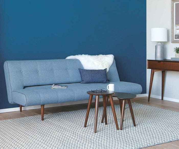 A modern mid-century living room features a blue sofa, the Juneau Nest Of Tables, a stylish rug, and a console table adorned with a lamp and decor. A striking blue accent wall contrasts beautifully with light flooring and neutral furnishings, resulting in an inviting and elegant living space.