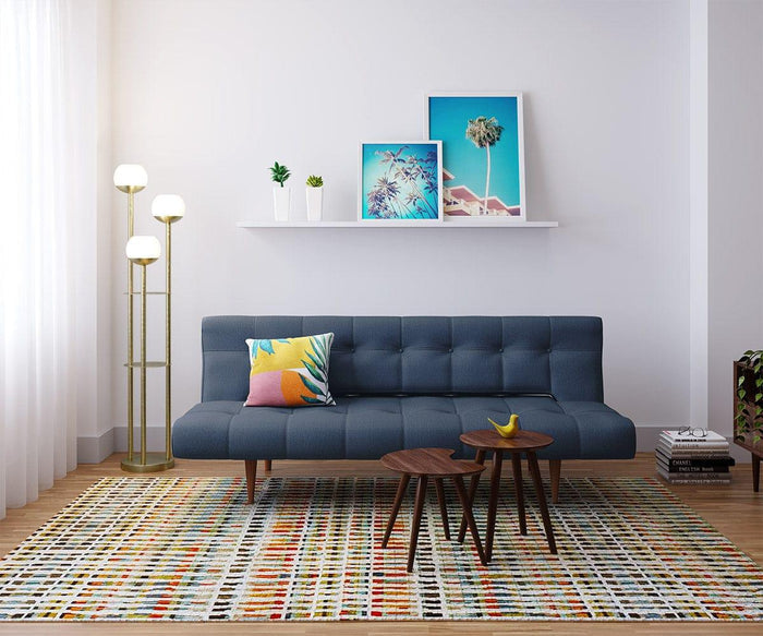 Modern living space with a blue sofa, colorful pillow, Juneau Nest Of Tables in a mid-century modern style, a banana, a floor lamp, shelf with plants, framed art, and a patterned rug on wood flooring. Bright natural light enters from a window with sheer curtains.