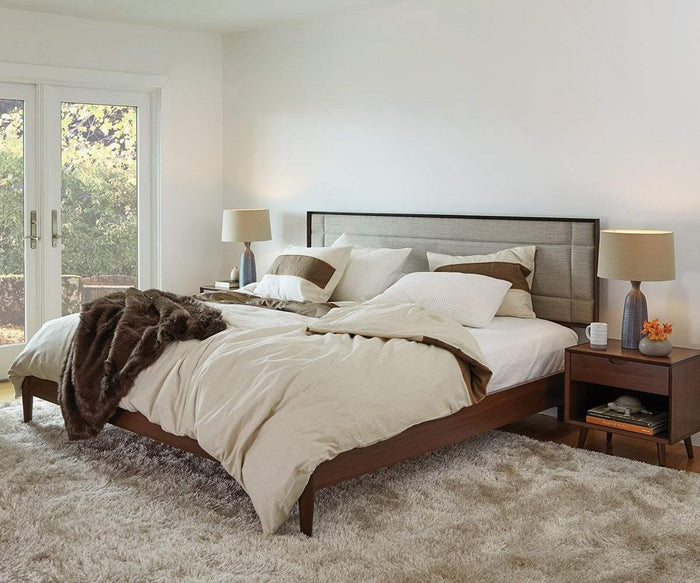 A cozy bedroom with a wooden bed, beige bedding, and a fluffy rug. The space features two Juneau Nightstands with lamps on either side of the bed, complementing the overall bedroom furniture. Sunlight streams through glass doors, illuminating the room, while a brown blanket is draped over the bed.