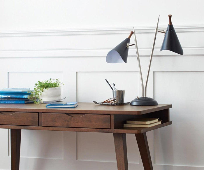 A wooden desk with books, a potted plant, a notebook, a pen holder, and the sleek Kabrino Table Lamp with adjustable shades is set against a white paneled wall.