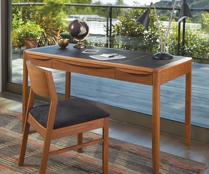 A wooden desk and chair sit by large windows with a view of greenery. On the desk are a globe, two Kabrino Table Lamps with adjustable shades, a phone, a notebook, and a pen. A patterned rug is on the floor beneath the desk.