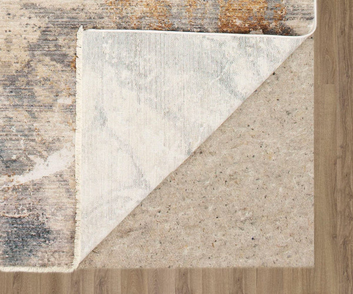 A corner of a patterned rug is lifted, revealing a beige non-slip rug pad underneath on a wooden floor—perfect for showcasing your durable area rug or the stylish Karastan Echo Tesoro Rug - Cream with its abstract motif.