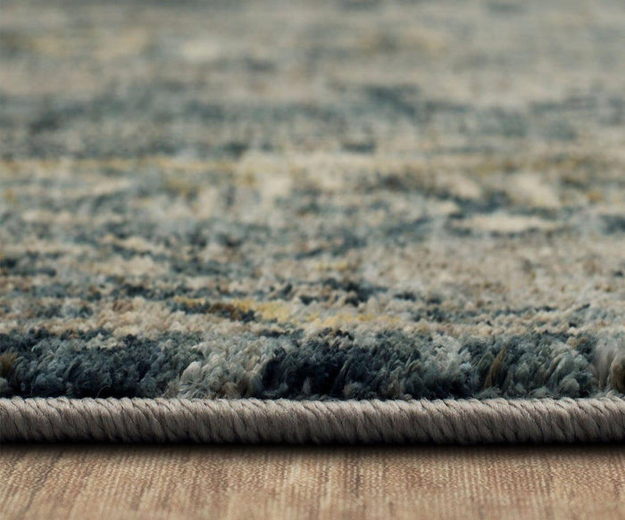 Close-up side view of the Karastan Marash Karamar Rug in Blue, showcasing its textured surface with gray and beige accents as it lies flat on a wooden floor. The rug’s edge and plush fibers are sharply focused, emphasizing its cozy appeal.