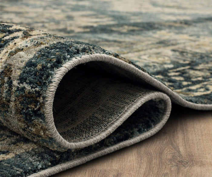 A close-up of the Karastan Marash Karamar Rug - Blue showcases its textured, soft surface in blue, beige, and green tones, partially folded over on a light wooden floor to reveal its woven underside and stitched edge. This durable rug effortlessly combines comfort and style for any space.