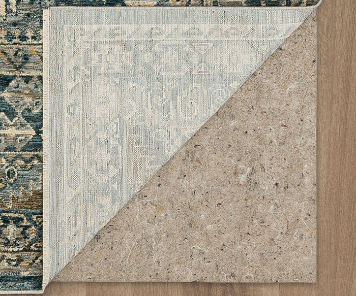 A corner of the Karastan Marash Karamar Rug in blue is lifted to reveal a durable felt rug pad underneath on a wooden floor.