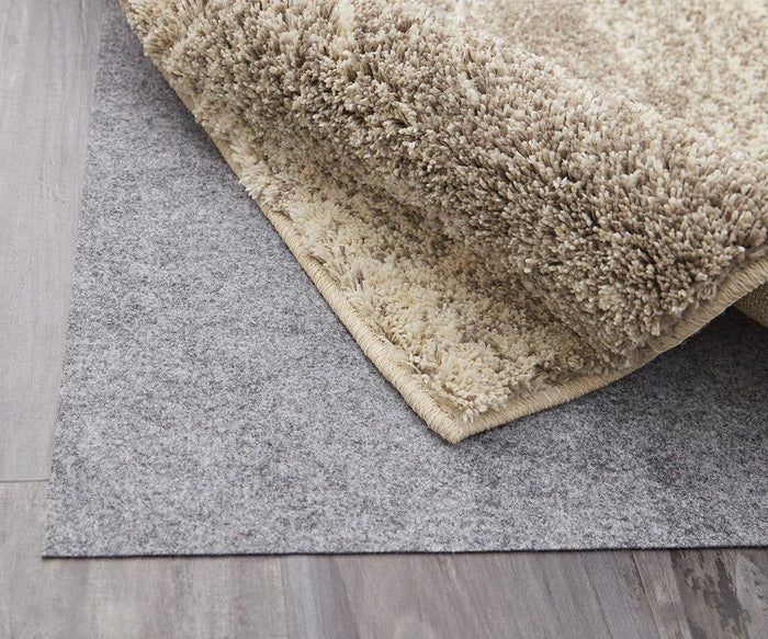 A close-up of a beige shaggy rug corner flipped over, revealing a gray Karastan Rug Pad underneath, both lying on a wooden floor.