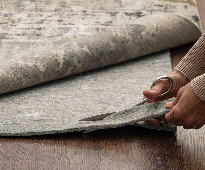 A person uses scissors to cut a Karastan Rug Pad on a wooden floor while an area rug is partially rolled up in the background, ensuring added protection for their space.
