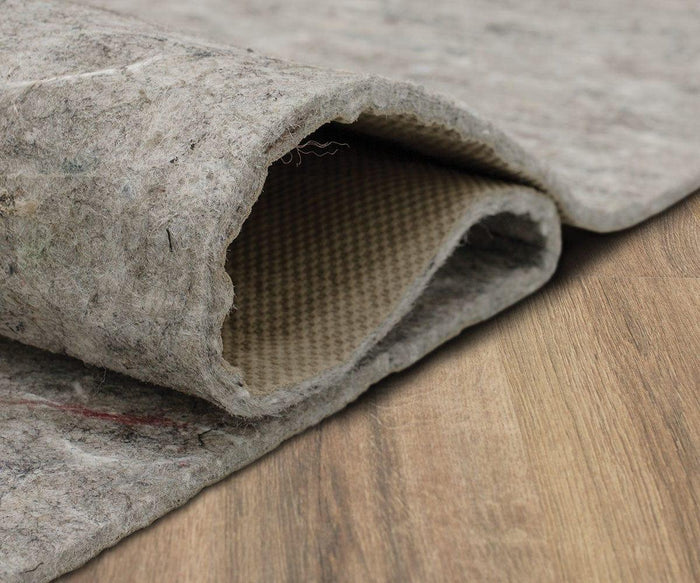 A close-up of the Karastan Rug Pad in gray, featuring a fabric texture and non-slip design, is shown partially folded to reveal its underside while lying on a wooden floor to provide added rug protection.