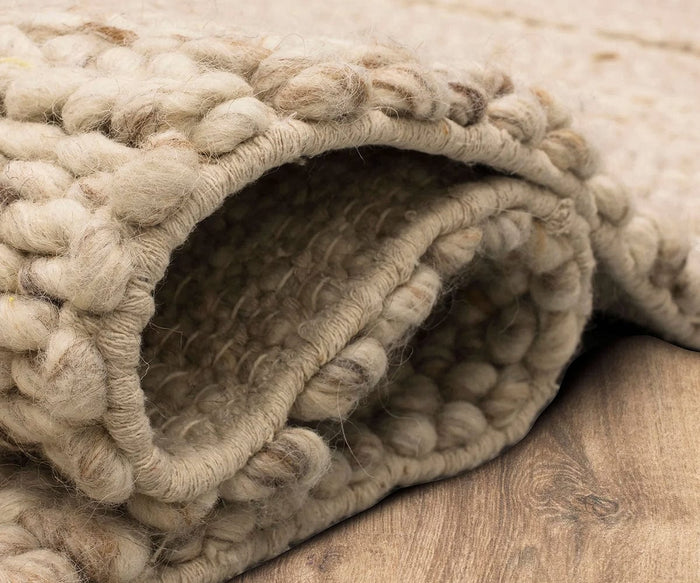 A close-up of the Karastan Tableau Roma Rug in Oyster, featuring a thick, hand-loomed wool weave with chunky texture, partially rolled on a wooden floor.