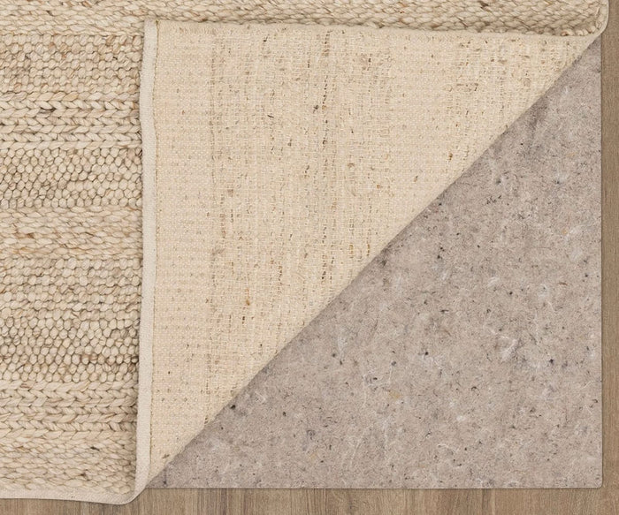 A Karastan Tableau Roma Rug in Oyster is folded back at one corner, revealing a tan felt rug pad underneath on a wood floor.
