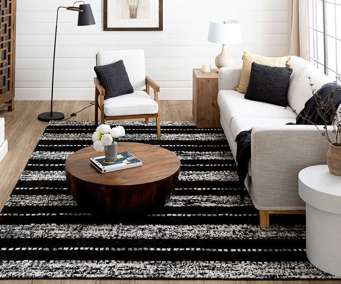 Modern living room with a white sofa, wooden armchair, round wooden coffee table, and the Karastan Traverse Lanes Rug in Charcoal featuring a striking black-and-white stripe pattern—crafted from durable polyester and perfectly suited for high-traffic areas—complemented by a floor lamp and decor including pillows, books, and a small vase of flowers.