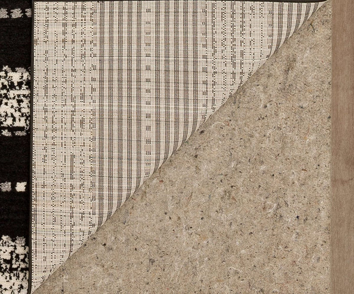 A corner of the Karastan Traverse Lanes Rug - Charcoal is flipped over, revealing its textured backing. This modern rug features a black and white geometric design, while the underside is light tan with visible fibers—a durable polyester rug perfect for any high-traffic area.