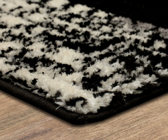 Close-up of the corner of the Karastan Traverse Lanes Rug in Charcoal, featuring a striking black and white pattern on a wooden floor. The image highlights the rug’s soft texture and stitched edges. Crafted from durable polyester, this rug is perfect for high-traffic areas.