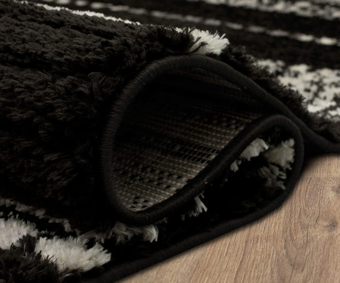 A close-up of the Karastan Traverse Lanes Rug in Charcoal, partially rolled on a wooden floor, showcases its black and white textured pattern, thick and soft fibers, and woven underside—making it an ideal choice for high-traffic areas thanks to its durable polyester construction.