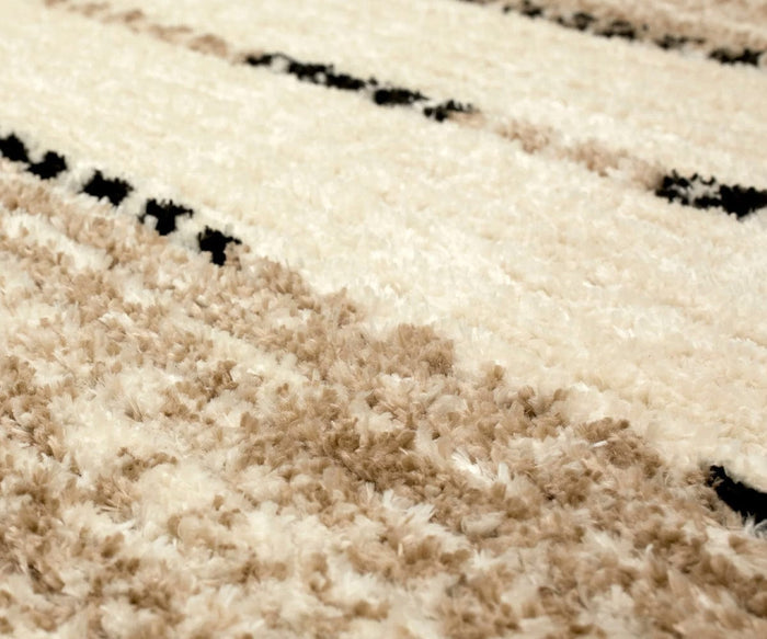 Close-up of a soft, fluffy area rug with beige, cream, and black abstract patterns. Crafted from durable polyester, the texture appears plush and inviting with subtle color variations throughout—much like the Karastan Traverse Lanes Rug in Tan.