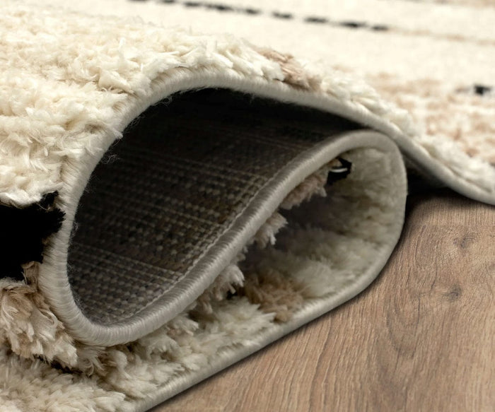 A close-up of the Karastan Traverse Lanes Rug - Tan, a beige, fluffy and durable polyester rug with the edge curled up to reveal its woven underside, lying on a wooden floor.