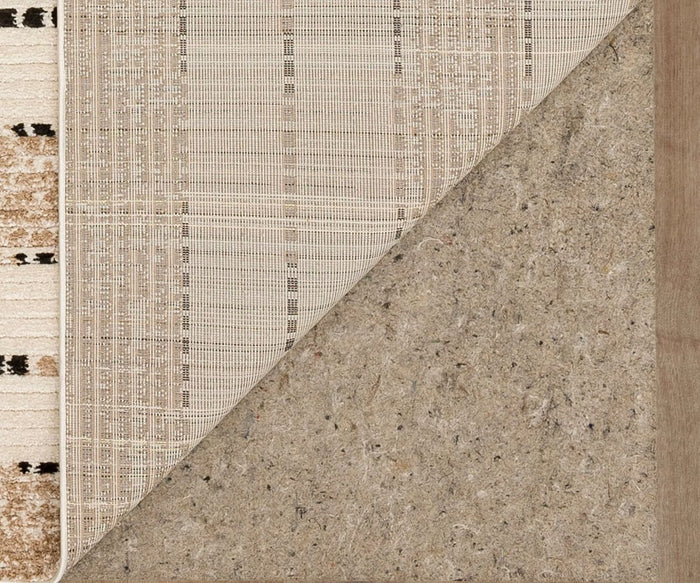 A close-up of a Karastan Traverse Lanes Rug - Tan corner flipped over reveals three distinct layers: a patterned top made from durable polyester, a mesh-like middle layer, and a thick, felt-like bottom layer—all displayed on a brownish wooden floor in signature Bobby Berk style.