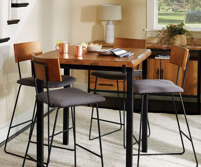 A wooden dining table with four chairs, copper mugs, a plate with sliced lime, and magazines on top. The Karsten Bar Stool adds a touch of natural charm beside the table. In the background, a lamp, plant, and cabinet are visible as sunlight streams through the window.