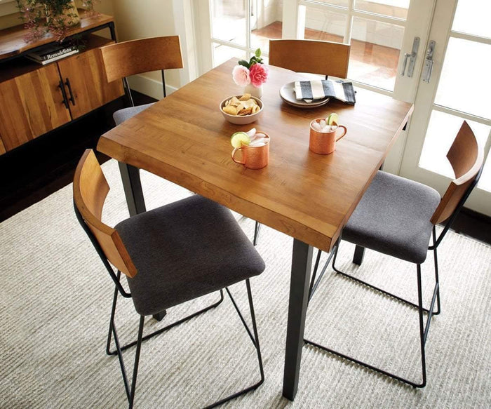 A wooden dining table with four gray cushioned chairs, three copper mugs with drinks, a plate of cookies, a bowl of crackers, and a vase of flowers sits in a bright room with double doors. Beside the table stands the Karsten Bar Stool, providing extra seating and perfectly complementing the inviting atmosphere.