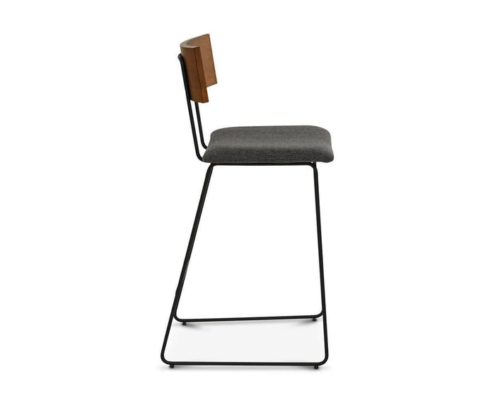 Side view of the Karsten Bar Stool, featuring a modern industrial design with a black metal sled-style frame, dark gray cushioned seat, wooden backrest, and horizontal support bar connecting the legs near the base.