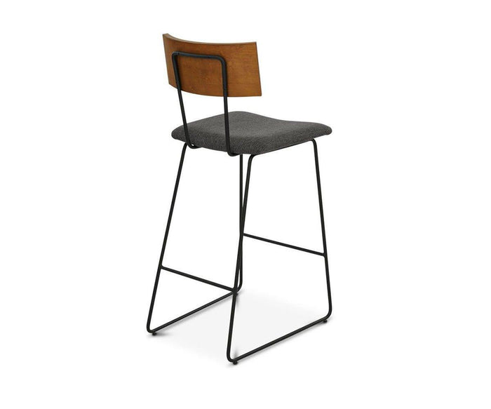 The Karsten Bar Stool features a modern industrial design with a black metal frame, a dark grey upholstered seat, and a wooden backrest, displayed at an angle against a white background.