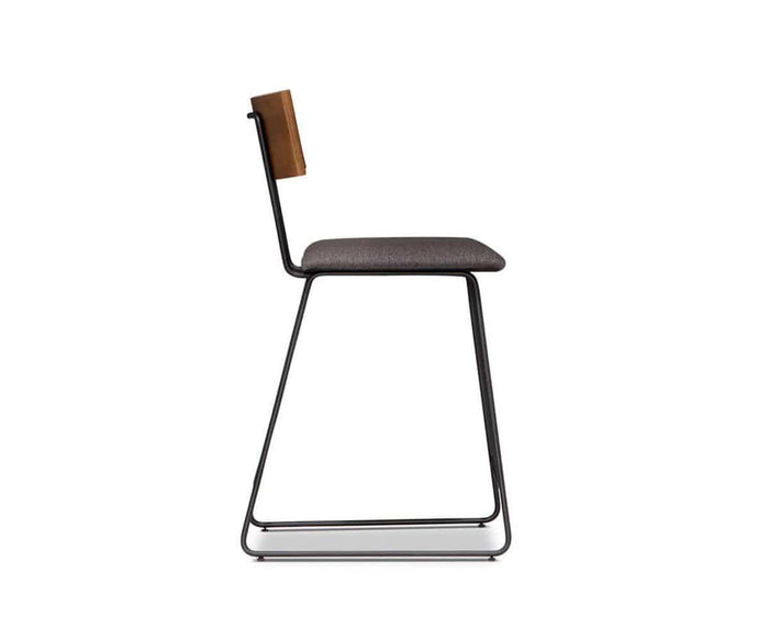 The Karsten Counter Stool with Back features a black metal frame, gray cushioned seat, and wooden backrest in a style reminiscent of a rustic industrial stool. It is shown in profile against a white background.