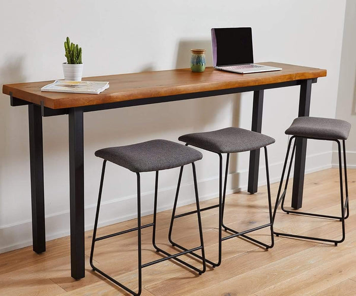 A long wooden table with black metal legs has three Karsten Counter Stools—gray, cushioned seats in a rustic industrial style—tucked underneath. On the table are a small potted cactus, a closed laptop, a mug, and a magazine. The scene is set on a light wood floor.
