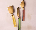 Three gold cheese knives featuring borosilicate glass handles in translucent multi-colored shades are arranged on a light beige surface. This exquisite Kota Cheese Set is beautifully handcrafted in India, adding a unique touch to your table.