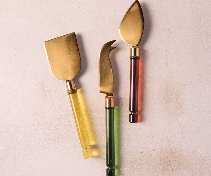 Three gold cheese knives featuring borosilicate glass handles in translucent multi-colored shades are arranged on a light beige surface. This exquisite Kota Cheese Set is beautifully handcrafted in India, adding a unique touch to your table.