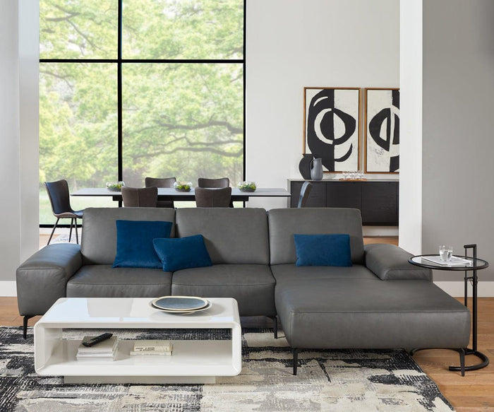 A contemporary living room features a gray sectional sofa adorned with blue accent pillows, a white coffee table, and the Krom C-Table—with its adjustable height—positioned next to the seating area. Abstract artwork decorates the wall, and expansive windows reveal views of trees outside. In the background, a dining area with six chairs completes the space.