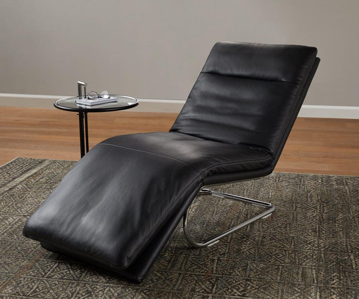 A modern black leather chaise lounge with a metal base sits on a patterned rug beside the Krom C-Table—an elegant, modern side table displaying eyeglasses and a drink—against a plain wall and wooden floor.