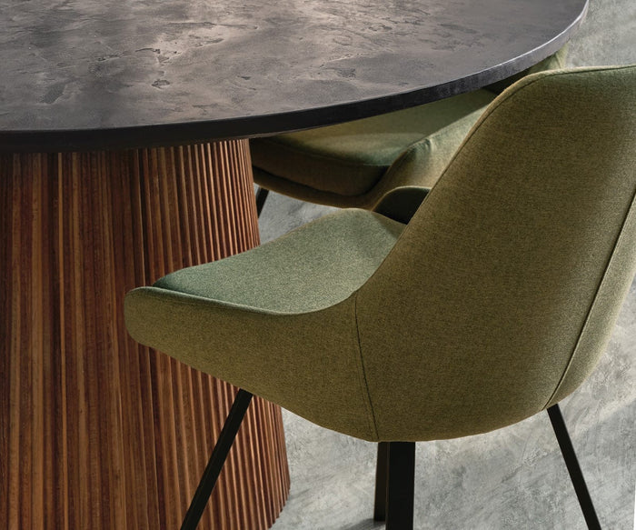 Certainly! Here’s the sentence rewritten using your product data: A close-up of two Kyla Swivel Dining Chairs with green upholstery, cushioned seats, and black legs featuring metal supports, placed beside a round table with a textured dark surface and a vertical wooden slat base.